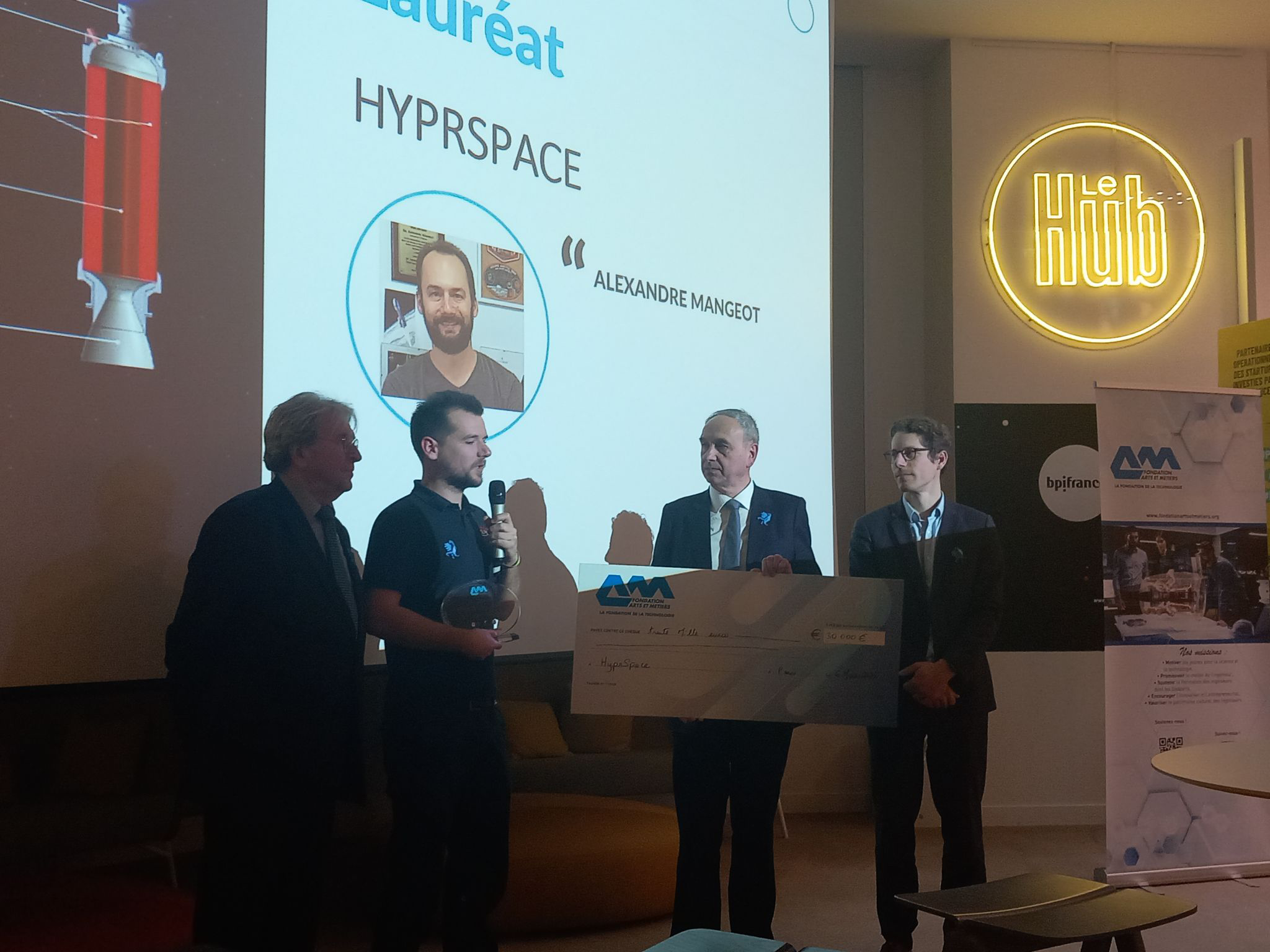 HyPrSpace founder receiving an award during the Grand Prix A&M ceremony. The image shows the presentation of a symbolic check and a trophy in front of an audience at Le Hub by Bpifrance.
