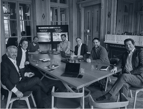 A group of investors and team members seated around a large meeting table with open laptops, documents, and a screen displaying a rocket. Professional setting in a historic room.