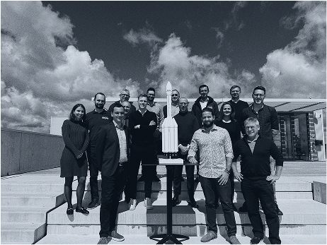HyPrSpace team standing proudly behind a scale model of their rocket on outdoor steps, under a blue sky with clouds. A symbolic image of ambition and team unity.