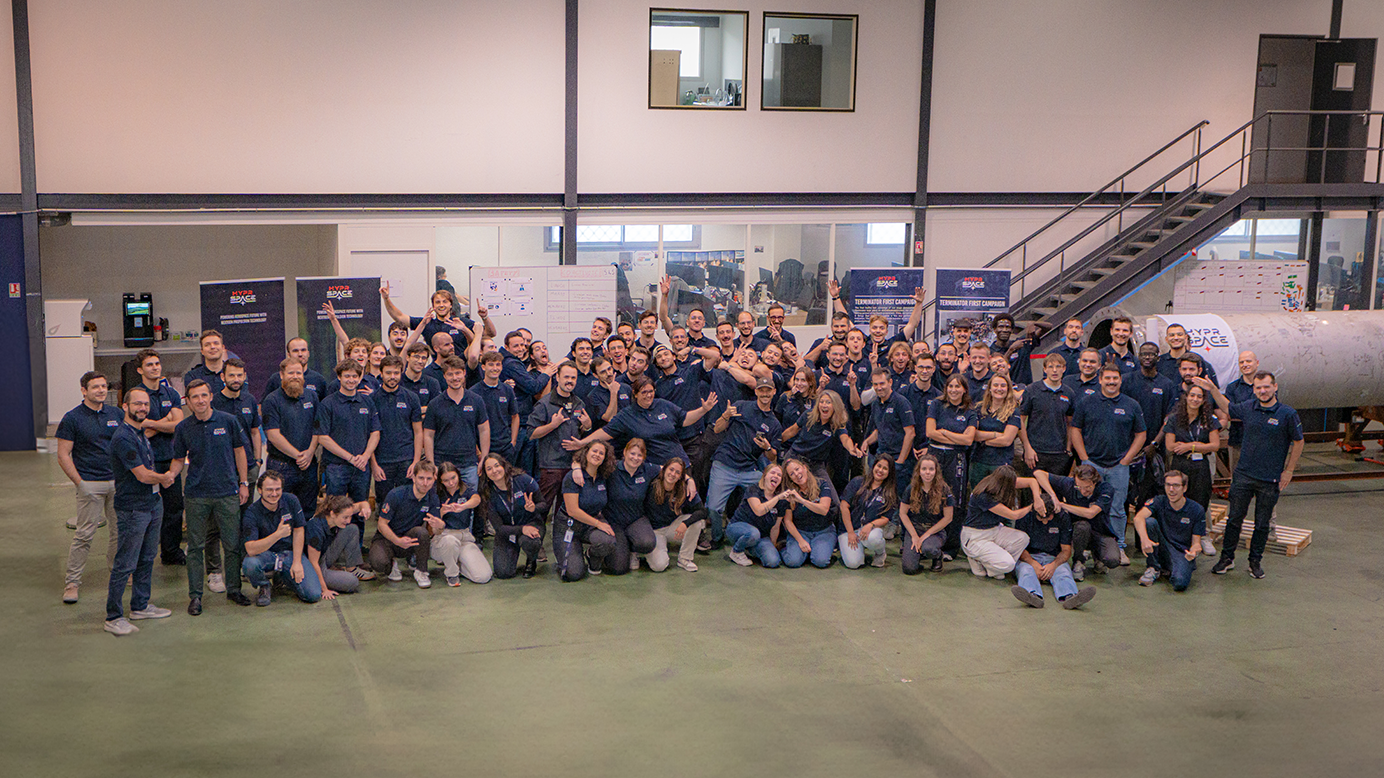 Large group photo of the HyPrSpace team in their workshop, smiling, gesturing playfully, and posing in front of propulsion equipment. A strong expression of company culture.