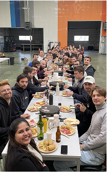 Dozens of HyPrSpace team members sharing a meal at a long table in a large industrial hangar, showcasing the team spirit and camaraderie of the company.
